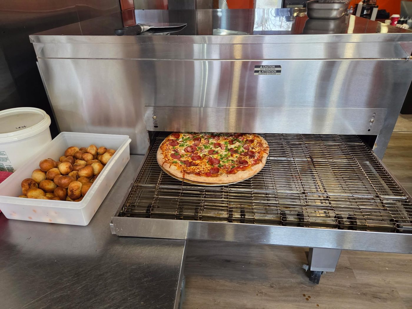 A fresh pepperoni and bacon pizza coming off the conveyor oven at Old School Pizza, with a tray of fresh dough balls beside it.